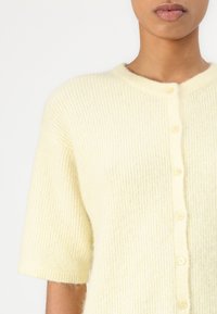 Kremfarget kortermet cardigan i ribbestruktur, med rund hals og seks knapper foran. Mykt, lodden materiale.