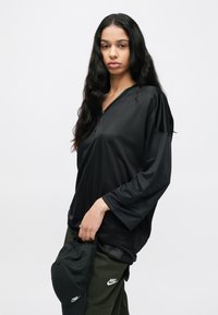 Μαύρο πουκάμισο με μακριά μανίκια, ριγέ υφή και V-λαιμόκοψη, συνδυασμένο με παντελόνι λαδί. Μια μαύρη καπέλο κρέμεται από τη ζώνη.