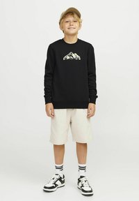 Felpa nera con grafica di montagna, pantaloni corti beige, sneakers bianche con dettagli neri e un cappellino chiaro. Fit semplice e rilassato, in materiale di cotone.