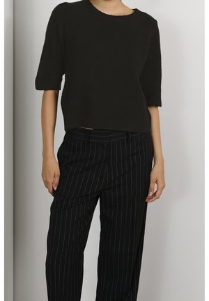 Osoba ubrana w czarny, krótki sweter z krótkim rękawem oraz czarne spodnie w cienkie paski, stojąca na jednolitym tle.