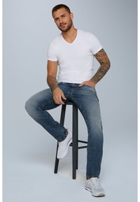 Camiseta blanca de cuello en V combinada con jeans azules desgastados, sentado en un taburete negro. Lleva zapatillas blancas, con brazos tatuados visibles. Fondo neutro.