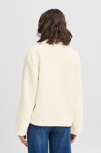 Lyscremet, tekstureret fleecejakke med afslappet pasform, sjalskrave, sænkede skuldre og lige kant. Båret over blå jeans.