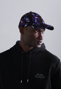 Gorra de béisbol con un patrón floral negro, con acentos en azul y rojo. Se lleva con una sudadera negra que presenta una marca sutil.