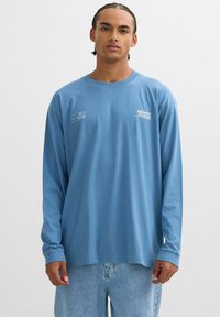 Camiseta de manga larga azul de algodón con un corte relajado, que presenta texto en blanco y acentos gráficos en el pecho. La lleva con unos vaqueros de mezclilla azul claros.