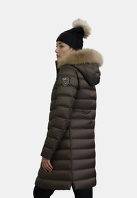 Pardessus marrone con cuciture orizzontali, cappuccio foderato in pelliccia e patch con logo sulla manica. Indossato con un berretto nero con pompon.