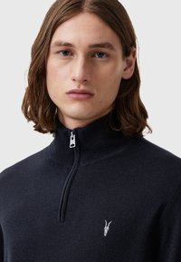 Navy zip-up Pullover aus weichem Strickstoff, mit hohem Kragen und kleinem weißen Emblem auf der Brust. Minimalistisches Design.
