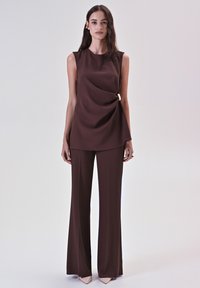 Donna che indossa un top drappeggiato senza maniche di colore marrone scuro con dettaglio a chiusura e pantaloni larghi coordinati, in piedi contro uno sfondo leggero e semplice.