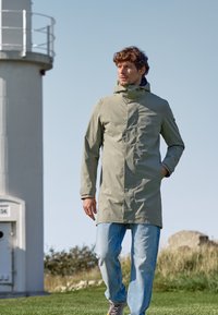Manteau imperméable gris clair avec une capuche, dotée d'une fermeture éclair avant et de poches latérales, associé à un jean bleu et des baskets grises.