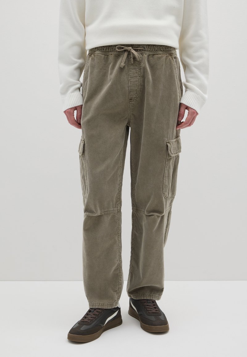 Licht olijfgroene corduroy cargobroek met een elastische tailleband, trekkoorddetail en twee zijzakken. Draagt met bruine sneakers.