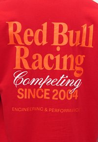 Κόκκινο πουκάμισο με πορτοκαλί κείμενο «Red Bull Racing», λευκό καλλιγραφικό «Competing», πορτοκαλί «Since 2004» και μικρό «Engineering & Performance» από κάτω.
