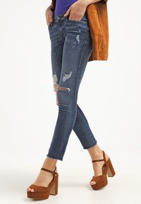 Jeans skinny de color azul oscuro con detalles desgastados y dobladillos deshilachados, combinados con tacones de plataforma marrones. El material parece vaquero.