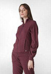 Mörkröd zip-up jacka tillverkad av mjukt material, med hög krage, sidofickor och elastiska ärmslut för en tät passform.