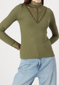Olijfgroene langemouwen top met geribbelde textuur, met kanten accenten bij de hals en mouwen, met een aansluitend ontwerp. Draag het met lichtblauwe jeans.