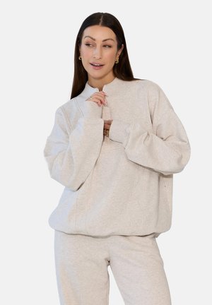 Kobieta o długich ciemnych włosach, ubrana w beżową, oversize'ową bluzę i dopasowane dresy, patrząca w lewo, z rękami blisko twarzy.