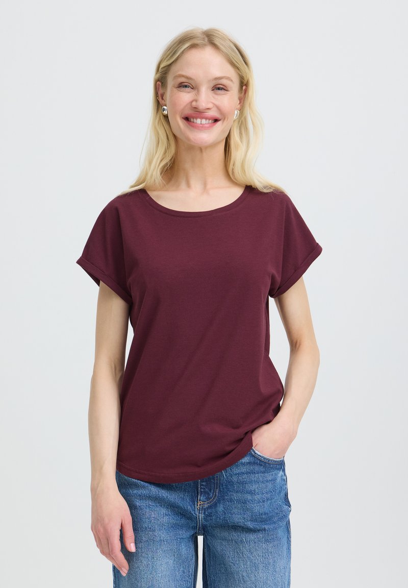 Femme souriante aux longs cheveux blonds porte des boucles d'oreilles argentées, un t-shirt bordeaux à manches courtes et un jean bleu, une main dans la poche, sur fond blanc.