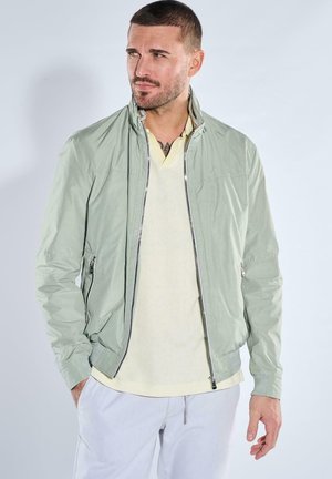 Mann trägt eine hellgrüne Bomberjacke über einem cremefarbenen Poloshirt und weißen Hosen, blickt seitlich vor einem schlichten Hintergrund.