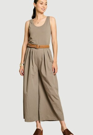 Vrouw draagt een mouwloze taupekleurige jumpsuit met een gevlochten bruine riem, armband, ketting en bruine open-toe platformschoenen.