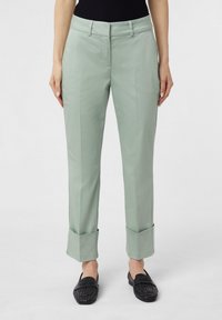 Jasnozielone dopasowane spodnie o prostym kroju, z mankietami u dołu i gładką fakturą, w zestawieniu z czarnymi plecionymi mokasynami.