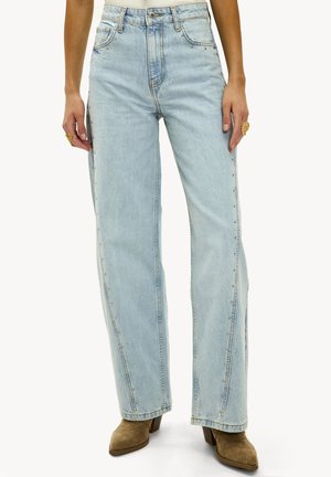 Lichtgewassen, wijde jeans met zijkantstudversieringen, gedragen met tan kleurige suede hoge hakken, gouden ringen en een witte top.