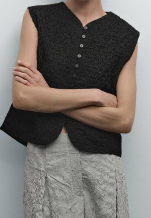 Person wearing a sleeveless black textured button-up top with vertical-striped light pants, arms crossed below the chest.