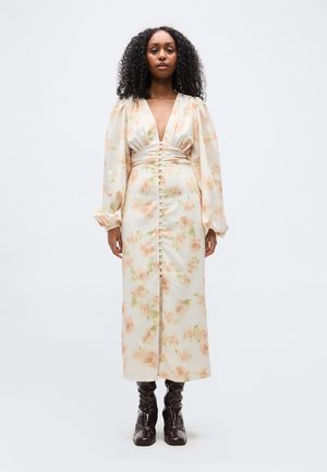 Žena v dlhých kvetinových šatách s nafúknutými rukávmi, gombíkovými detailmi na prednej strane a tmavohnedými čižmami do polovice lýtok, stojaca na bielom pozadí.