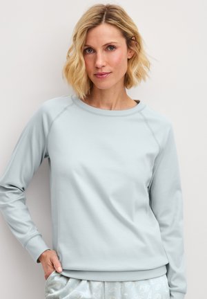 Blonde vrouw met lichtblauw sweatshirt met lange mouwen en lichtblauwe broek met patroon, staand tegen een effen achtergrond.