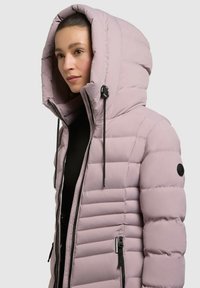 Chaqueta acolchada rosa con una gran capucha, bolsillos con cremallera y acentos en negro. Presenta un texto acolchado horizontal y cordón para ajuste.