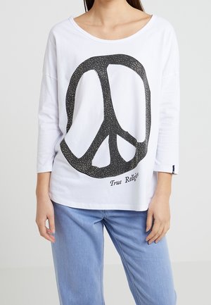 Femme portant une chemise blanche à manches longues avec un grand symbole de paix noir et le texte « "True Religion" », associée à un jean bleu clair.