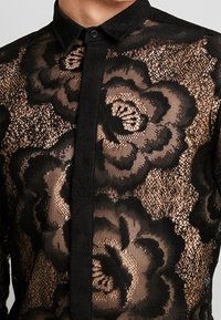 Camicia in pizzo nero con motivo floreale, trama trasparente e scollo con colletto. Presenta una chiusura frontale con bottoni e un bordo solido a contrasto.