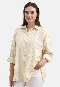 Camicia giallo chiaro con bottoni, caratterizzata da righe verticali viola, taglio rilassato, maniche lunghe arrotolate e colletto a punta.
