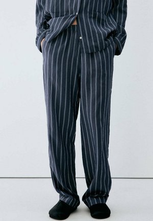 Personne portant un pyjama ample foncé à rayures blanches verticales et des chaussons noirs, debout les mains dans les poches devant un fond uni.