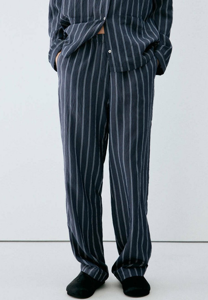 Personne portant un pyjama ample foncé à rayures blanches verticales et des chaussons noirs, debout les mains dans les poches devant un fond uni.