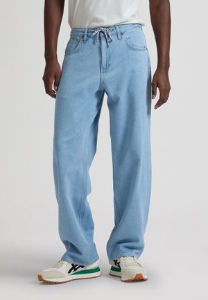 Mann trägt hellblaue locker sitzende Jeans mit Kordelgürtel und weißen Sneakers mit schwarzen und grünen Akzenten, steht vor schlichter Hintergrund.