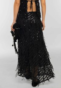 Falda larga negra con flecos que presenta un patrón diagonal, adornada con lentejuelas. Tejido texturizado, de largo máximo y diseño sin forro para superponer.