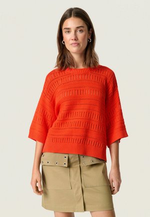 Heller orange gestrickter Pullover mit lockerer Passform, der strukturierte Streifen und einen gerippten Saum aufweist. Kombiniert mit einem beigen Rock mit Druckknopftaschen.