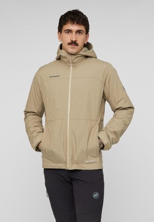 Mann mit Schnurrbart, der eine beige Kapuzenjacke und schwarze Hose trägt, steht mit den Händen in den Taschen vor einem einfarbigen Hintergrund.