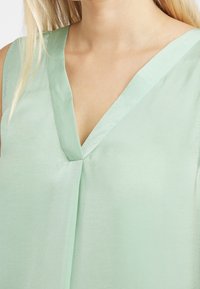 Blusa sin mangas de color verde claro con escote en V, hecha de una tela suave. Presenta un pliegue central y un diseño limpio y minimalista.
