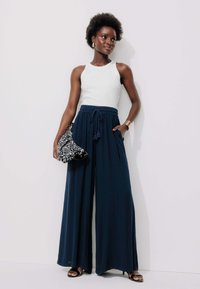 Pantalons larges bleu marine avec taille à cordon de serrage, associés à un débardeur blanc ajusté. Les accessoires comprennent une pochette noire et argentée et des sandales à talons.