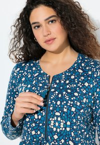 Top de estampado azul con cremallera en la parte delantera, escote redondo y una mezcla de formas abstractas en tonos blancos y azules más claros sobre una tela suave.