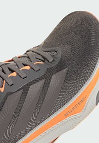 Scarpa sportiva grigia con una tomaia testurizzata, caratterizzata da dettagli arancioni e tre strisce scure. Suola testurizzata etichettata "DREAMSTRIKE+".