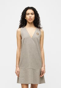 Γυναίκα με σγουρά μαλλιά που φοράει αμάνικο μπεζ καρό φόρεμα με V-λαιμόκοψη και χρυσό καρφίτσα μέλισσα σε απλό λευκό φόντο.