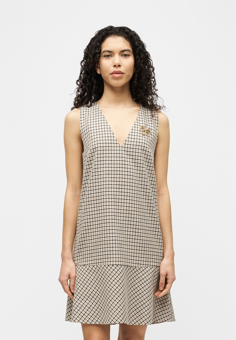 Γυναίκα με σγουρά μαλλιά που φοράει αμάνικο μπεζ καρό φόρεμα με V-λαιμόκοψη και χρυσό καρφίτσα μέλισσα σε απλό λευκό φόντο.