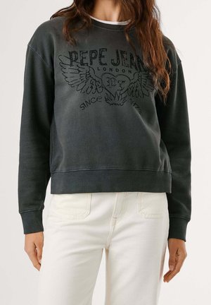 Vrouw draagt een vervaagde zwarte Pepe Jeans trui met een afbeelding van een gevleugeld hart en witte broek tegen een effen achtergrond.