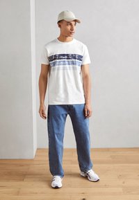 Weißes T-Shirt mit blauen horizontalen Streifen, kombiniert mit hellblauen Jeans und weißen Sneakers. Eine beigefarbene Baseballkappe rundet den Look ab.