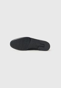 Semelle de chaussure noire avec un motif de bande de roulement nervuré à l'avant-pied et une section de talon lisse et légèrement surélevée, sur un fond uni.