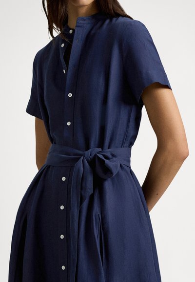 Kratka haljina tamnoplave boje s mandarin ovratnikom, prednjim gumbima i vezivanjem u struku. Izrađena od lagane tkanine s glatkom teksturom.
