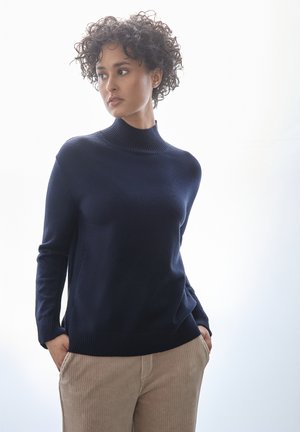 Pull à col roulé bleu marine en tissu tricoté doux, avec des poignets et un ourlet côtelés, associé à un pantalon en velours côtelé beige clair.