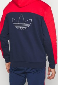 Marinblå och röd huvtröja med en vit Adidas-logotyp på baksidan. Tillverkad av mjukt tyg med en sportig design och ribbade muddar.