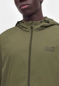 Gros plan d'une personne portant une veste zippée vert olive avec capuche et logo EA7 Emporio Armani sur la poitrine.