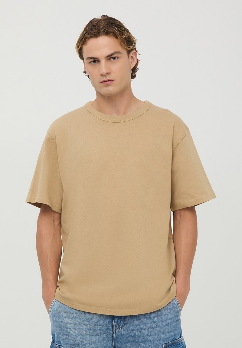 T-shirt in cotone beige con vestibilità rilassata, scollatura rotonda e maniche corte. Texture liscia, design minimale, senza motivi visibili.